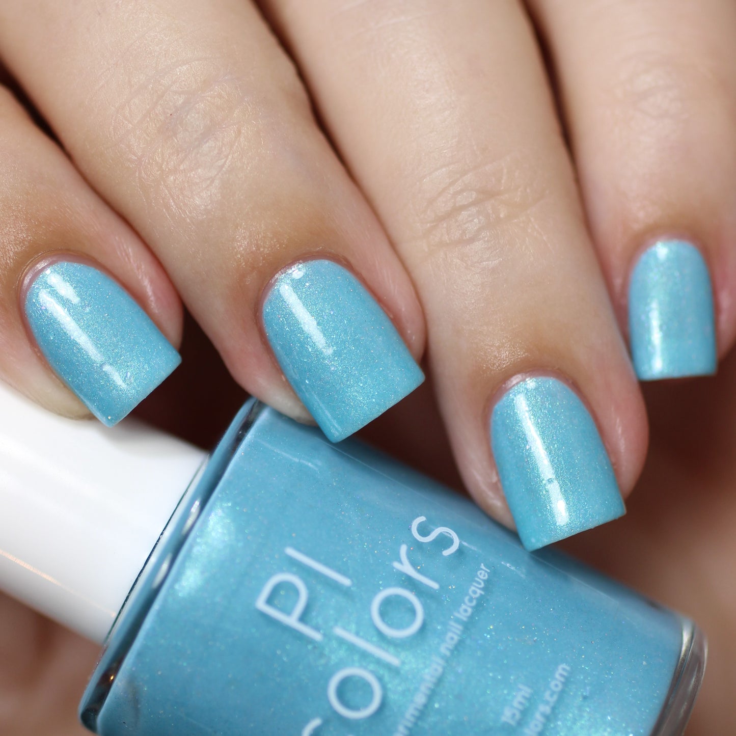 Close-up of a hand with manicured nails painted in the shimmering light blue shade, Berry Mousse.001 Blue Nail Polish. A bottle labeled "PI Colors" is placed nearby, showcasing its matching 10-free formula, while subtle hints of holographic powder add a dazzling effect.