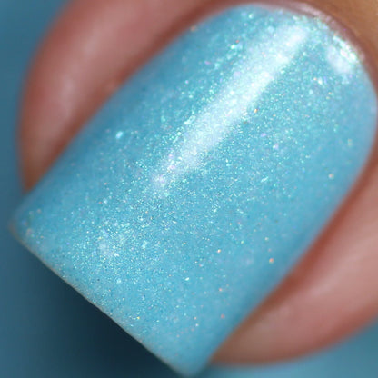 A close-up of a fingernail painted with PI Colors' Berry Mousse.001 Blue Nail Polish, enhanced by holographic powder, showcasing fine glitter particles that catch the light. The softly blurred background reflects a similar blue hue, creating an ethereal effect.