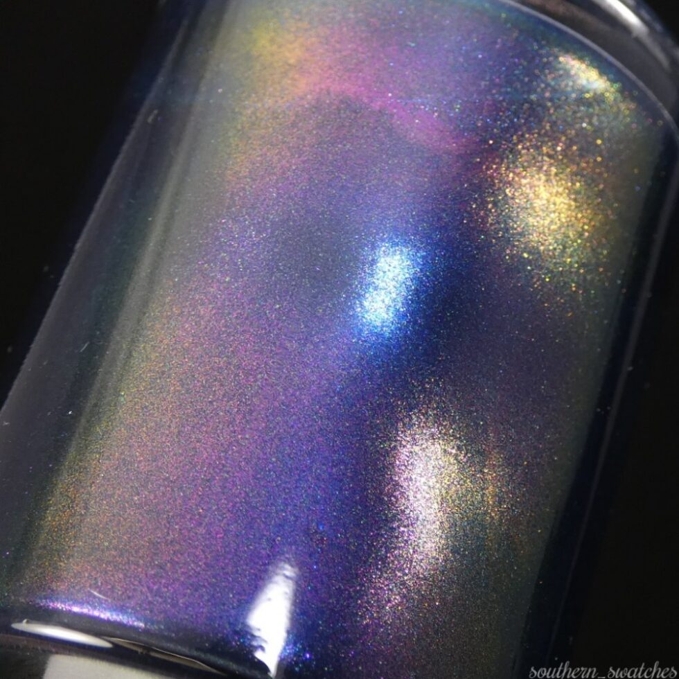 A close-up of PI Colors' "Judge None Choose One.000" luxury nail lacquer bottle showcases an iridescent finish that displays a vibrant spectrum of colors, including purple, blue, and gold. This multichrome nail polish reflects light with a smooth, glossy texture reminiscent of 90s nostalgia.