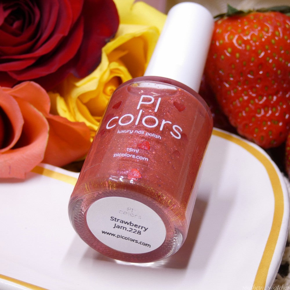 A bottle of PI Colors' Strawberry Jam.228 Red Nail Polish sits on a white surface. Surrounded by vibrant orange and yellow roses and a fresh strawberry, the shimmering pink-red shade with red glitter captures the essence of luxury effortlessly.