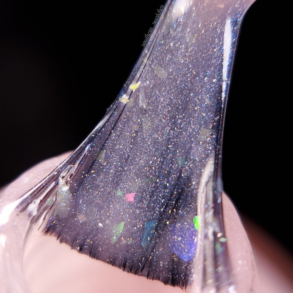 Close-up of a nail polish brush being pulled from a bottle of Special Week.000 Pink Nail Polish by PI Colors, showcasing clear, glittery luxury lacquer with iridescent flakes. The background is blurred to emphasize the sparkling texture and vibrant colors of this lavish polish.