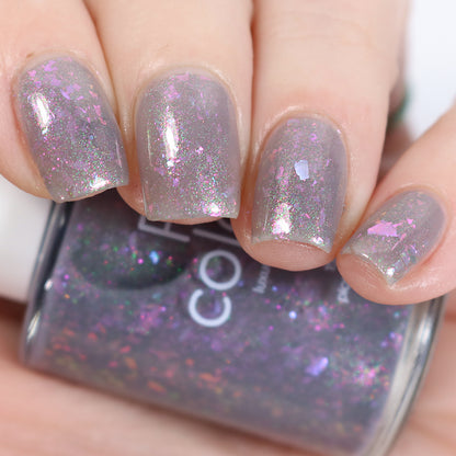 A close-up of fingernails showcases a glossy manicure using PI Colors' "The First Rule.000 Gray Nail Polish," which features a captivating mix of pink and purple iridescent flakies that catch the light beautifully, with the partially visible polish bottle in the background.