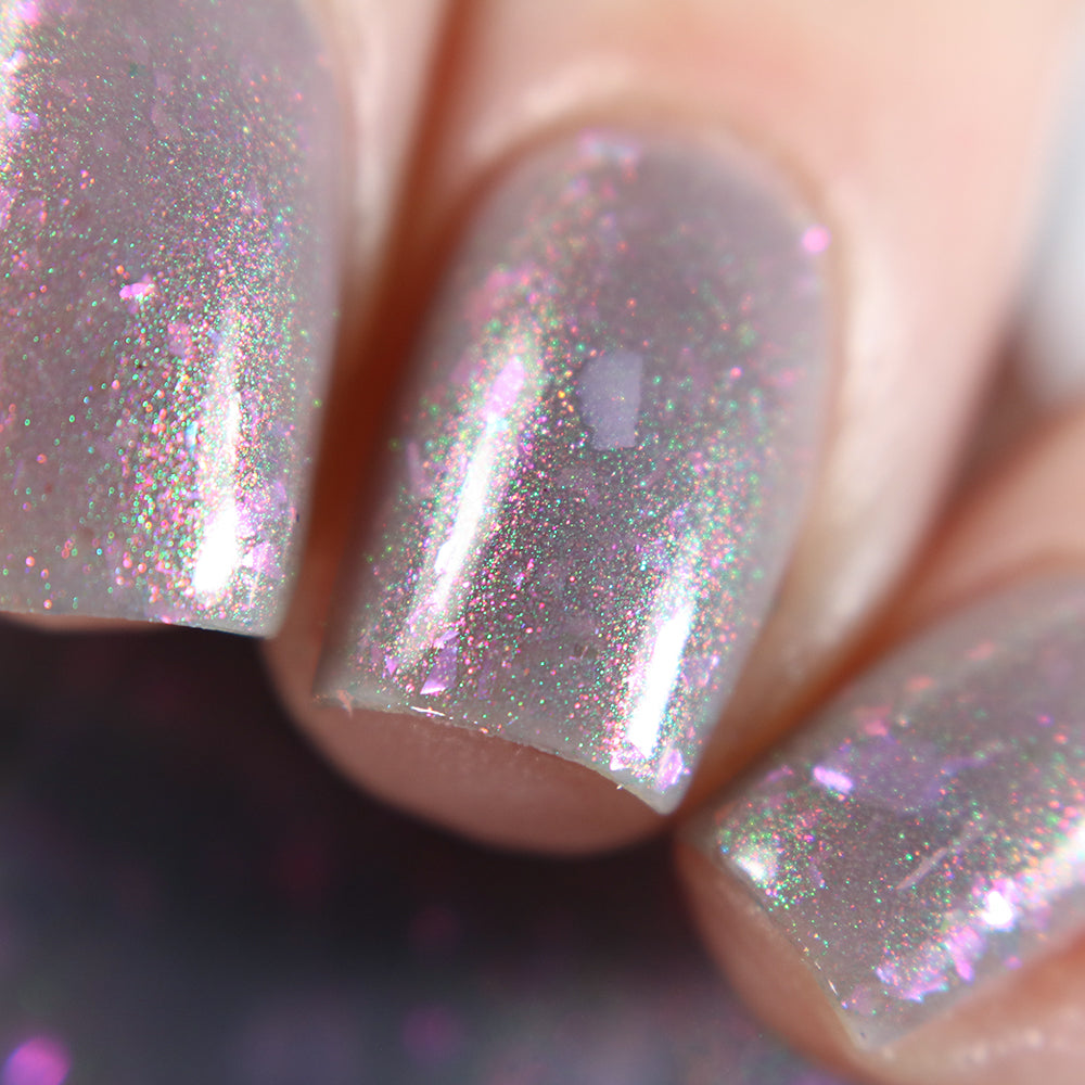 A close-up of nails showcasing PI Colors' "The First Rule.000 Gray Nail Polish," featuring a glossy, iridescent finish with mesmerizing flakies that reflect shades of pink, purple, and silver. The nail surface is smooth and shiny, capturing the light with a sparkling effect.