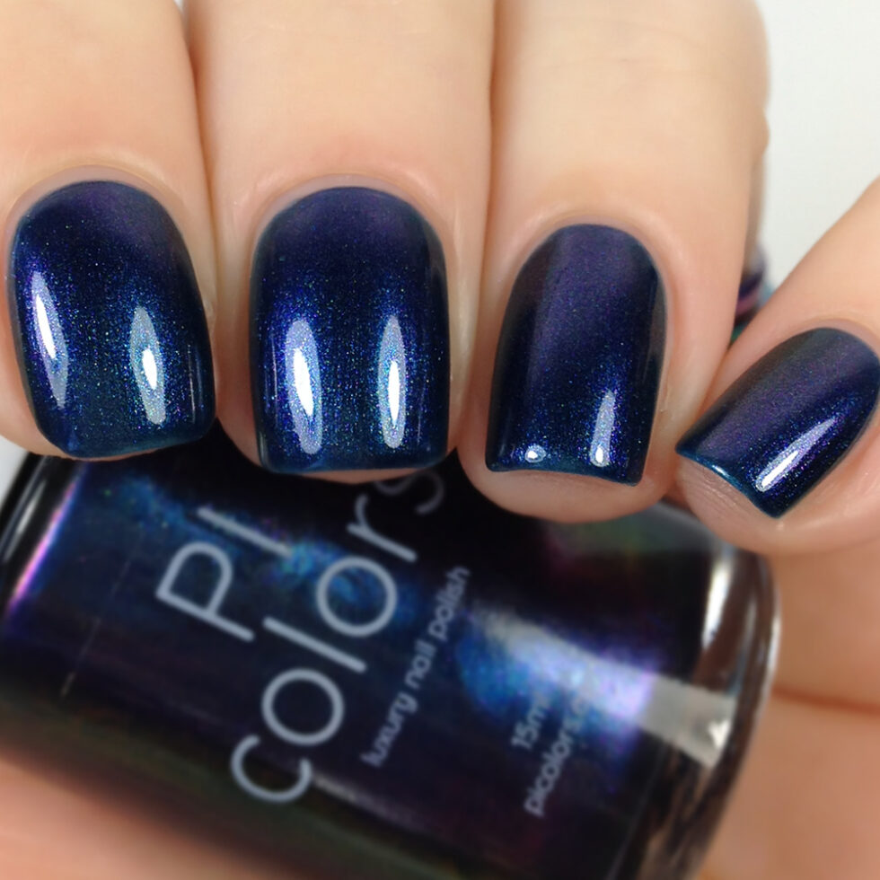 A close-up of a hand with neatly painted dark blue, shimmering nail polish evokes 90s nostalgia. The glossy, well-shaped nails are complemented by a partially visible bottle of PI Colors' luxury nail lacquer in the same rich hue, named Judge None Choose One.000, in the background.