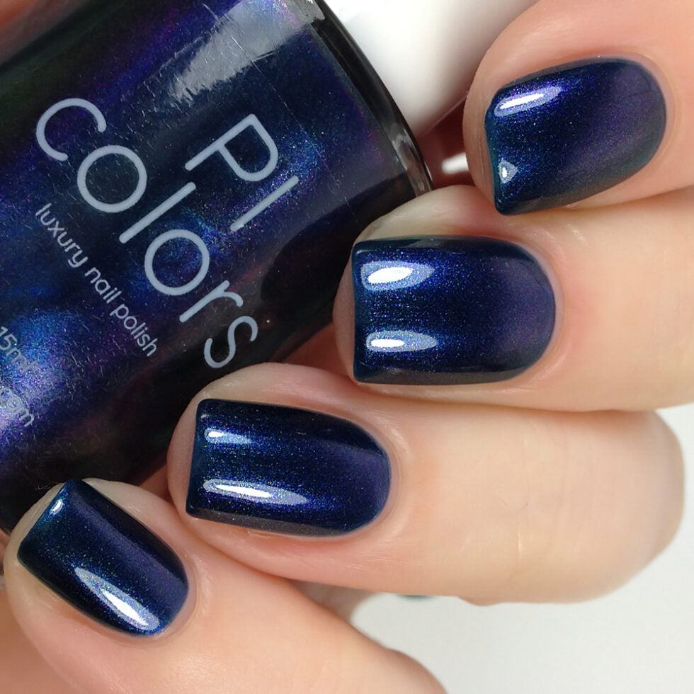 A hand with four neatly manicured nails painted in a glossy, deep blue nail polish brings back memories of 90s nostalgia. Held near the nails is a bottle labeled "PI Colors" featuring their product "Judge None Choose One.000," showcasing the same vibrant shade. The luxurious finish offers a metallic shine.