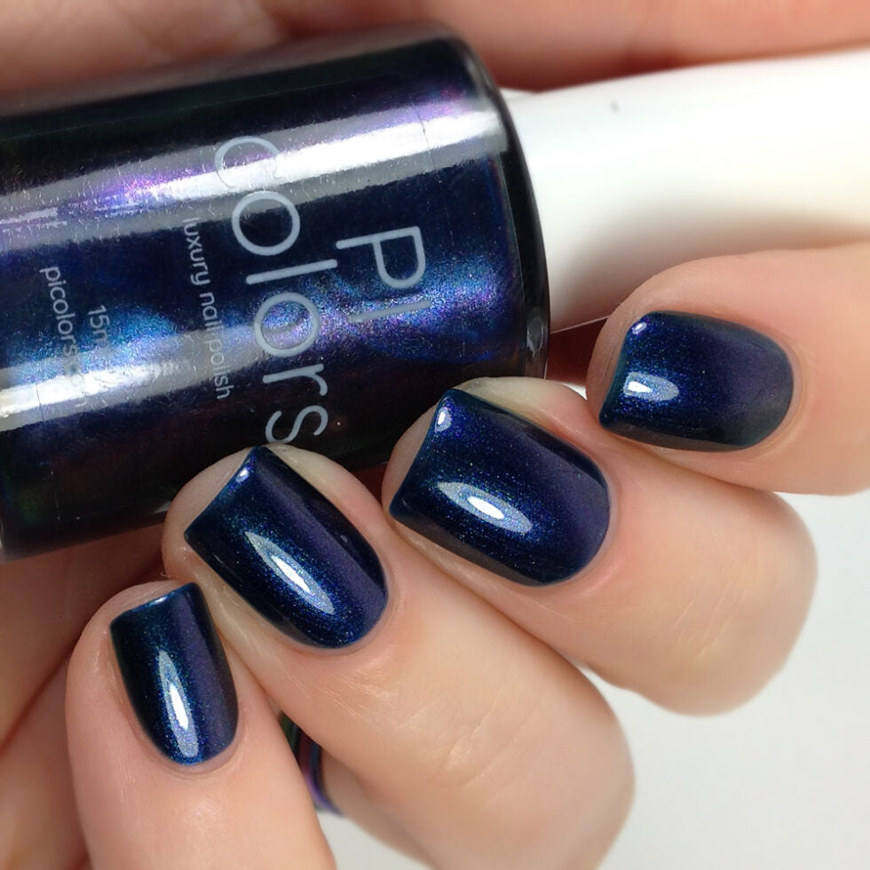 A close-up captures a hand with neatly manicured nails painted in PI Colors' glossily seductive "Judge None Choose One.000" dark blue nail polish, exuding 90s nostalgia. The shimmering effect enhances its allure, while a partially visible bottle of this luxury nail lacquer sits in the background, topped with a white cap.