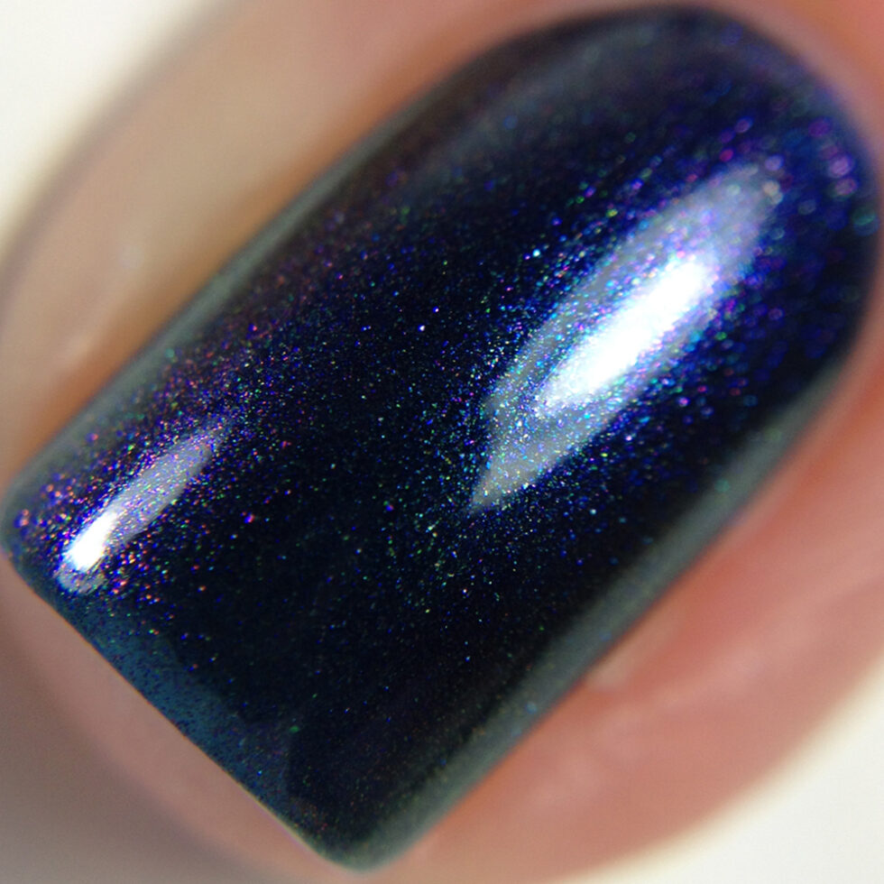 A close-up of a fingernail showcases PI Colors' Judge None Choose One.000, featuring luxurious nail lacquers in shimmering dark blue and purple multichrome hues. The glossy finish with subtle sparkles is beautifully highlighted against a soft, unfocused background, capturing the essence of 90s nostalgia.