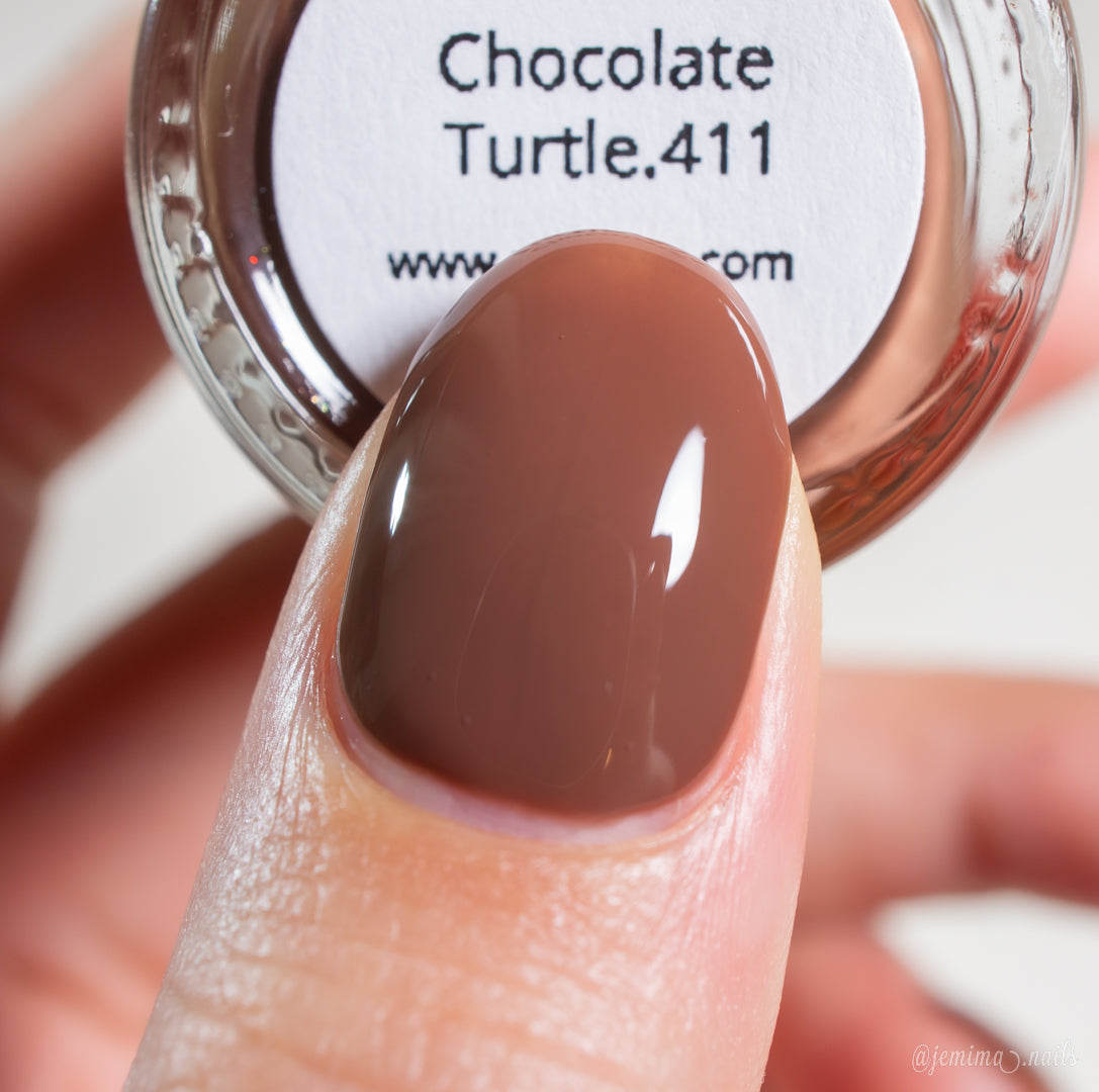 A close-up of a fingernail painted with PI Colors' Chocolate Turtle.411, a handmade nail polish with a glossy chocolate brown creme finish, held next to the matching bottle.