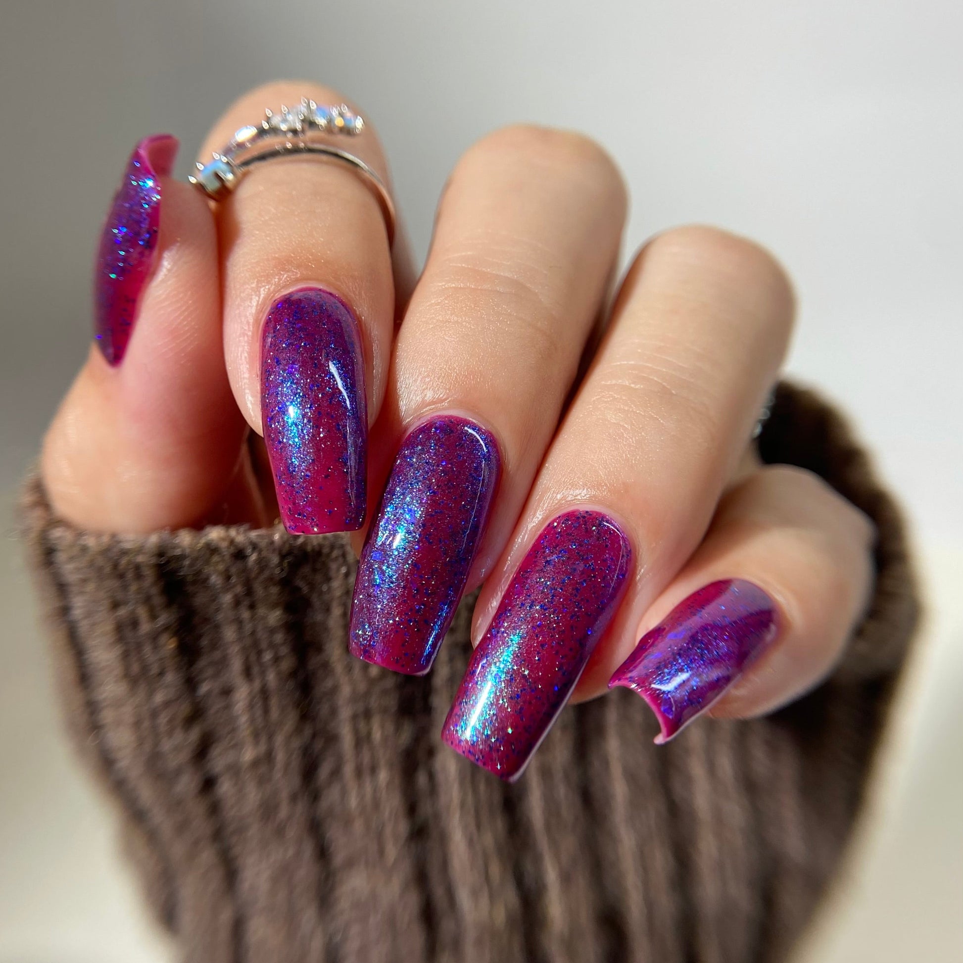 A close-up of a hand with long, almond-shaped nails painted in a shimmering purple and pink holographic glitter nail polish, wearing a brown knit sweater and silver rings.