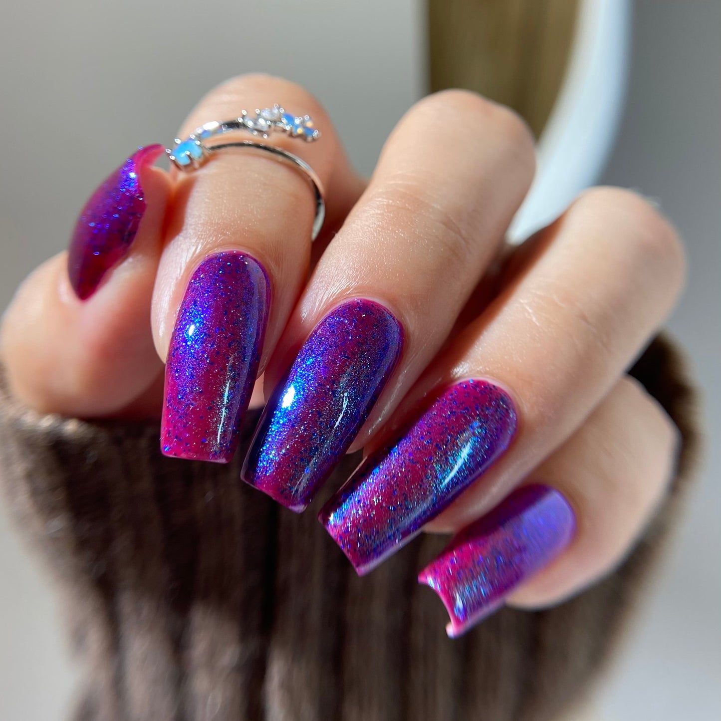 A close-up of a hand with long almond-shaped nails painted in a shimmering holographic purple and blue nail polish, wearing a silver ring with blue stones.