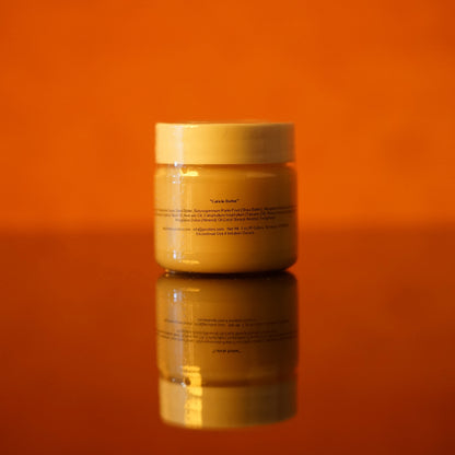 A jar of PI Colors Cuticle Butter, with its chocolate scent and orange hue, rests on a reflective surface against an orange background. The label, indicating a blend of organic butters, is subtly visible on the front of the jar.