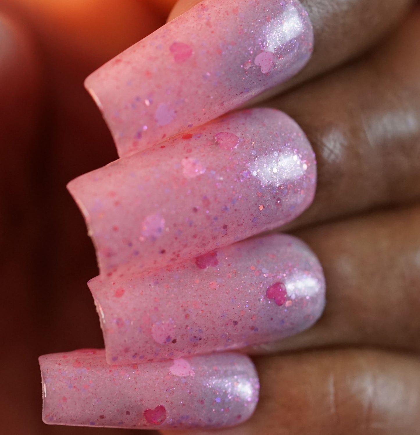 Close-up of fingers with long, glossy nails painted with PI Colors' "Pink Berry Heart.200," adorned with holographic hearts and showcasing a shiny, glittery texture and intricate nail art achieved using a 10-free formula.