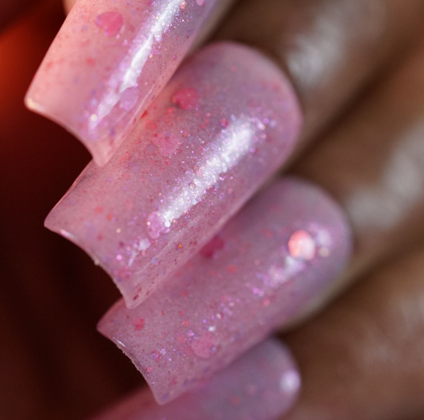 A close-up of nails with PI Colors' Pink Berry Heart.200 nail polish on a dark-skinned hand shows its pink glitter and holographic hearts over a translucent base, creating a sparkling effect. This product is 10-free for safe and stunning wear.