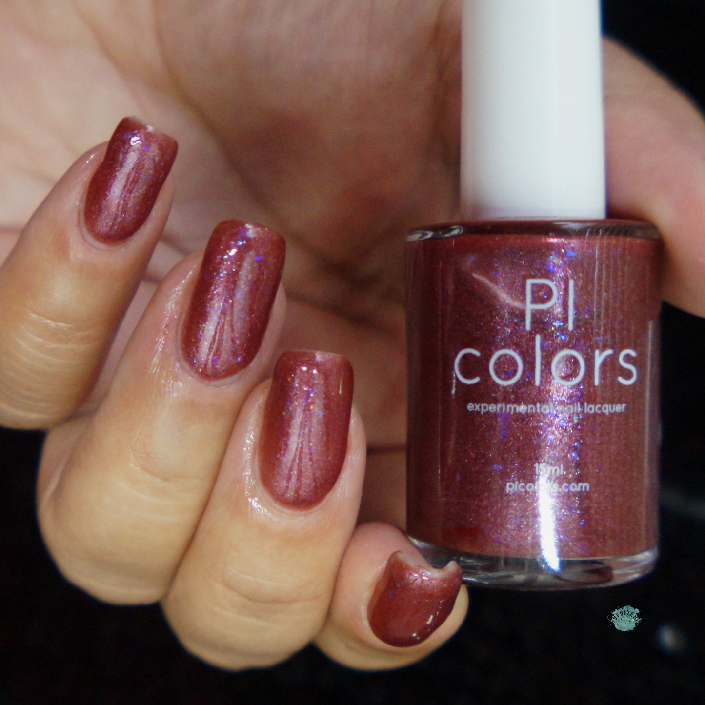 A hand displays nails painted with PI Colors' Quasar.156 Red Nail Polish, a luxury lacquer featuring a shimmering, reddish-brown hue. In the hand, there is a bottle of the same polish with its label clearly visible. The glossy finish showcases mesmerizing color-shifting flakies against a black backdrop.