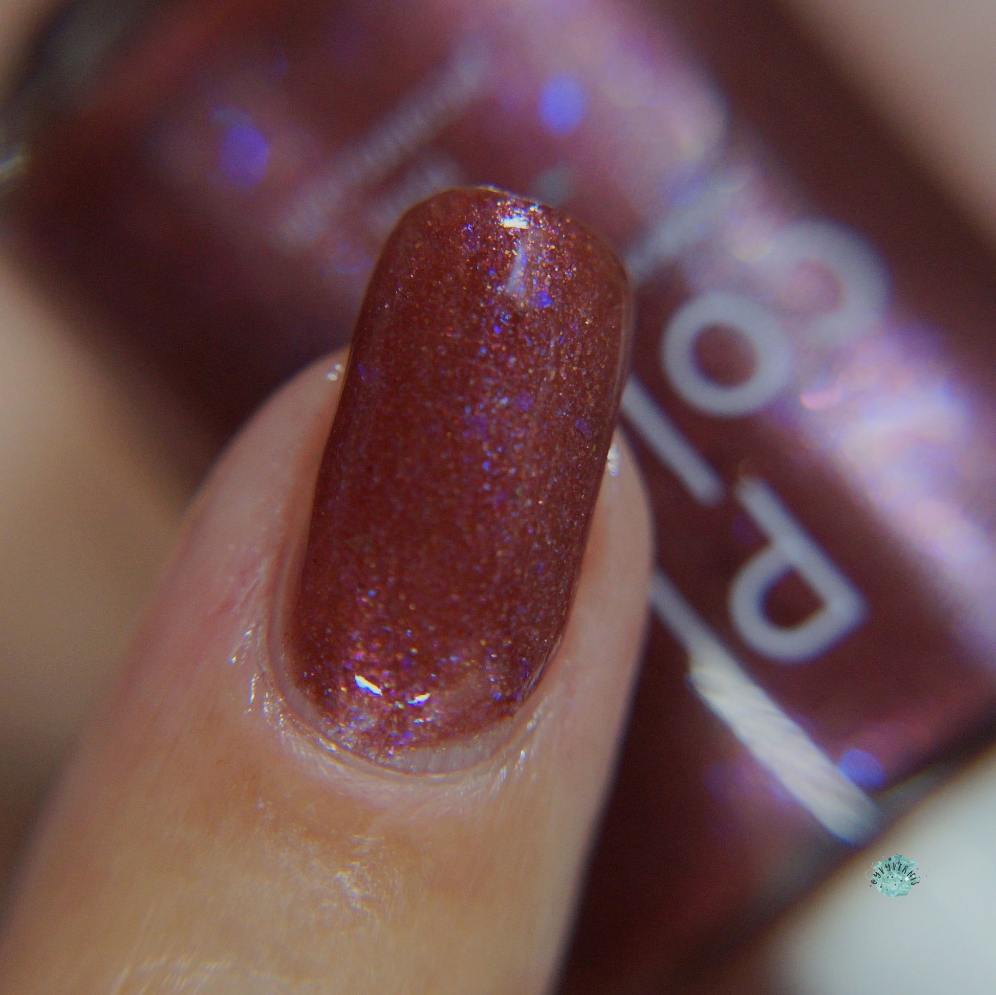 A close-up of a fingernail adorned with PI Colors' Quasar.156 Red Nail Polish showcases its shimmering reddish-brown luxury. In the slightly blurred background, the bottle hints at purple and blue shimmer that adds depth and dimension, creating a mesmerizing effect with its color-shifting flakes.