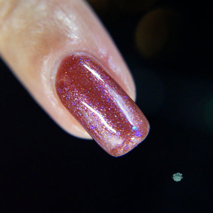 A close-up of a fingernail adorned with PI Colors' Quasar.156 Red Nail Polish, shimmering in maroon hues with tiny blue sparkles. The dark background accentuates the glossy finish and color-shifting flakies, highlighting the iridescence of this exquisite nail art.