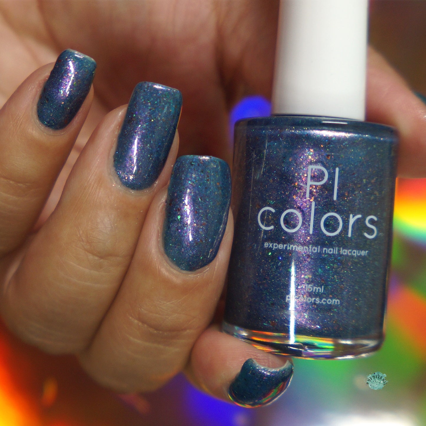 A hand with long nails painted in a shimmering deep blue hue holds a bottle of PI Colors' East of the Storm.029 Blue Nail Polish, highlighting its matching holographic shade enriched with color-shifting iridescent flakies. The background features blurred colorful light reflections.