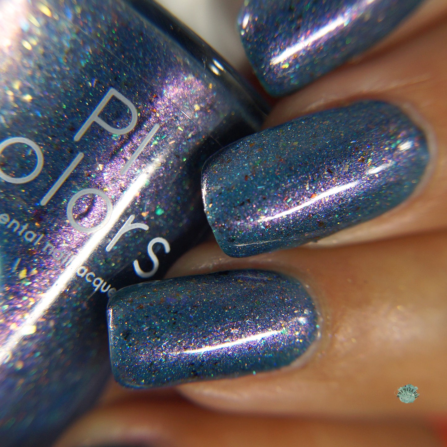 A close-up of fingernails showcases the shimmery East of the Storm.029 Blue Nail Polish by PI Colors, adorned with multicolored glitter. The glossy finish and iridescent flakies create a dazzling light-reflecting sparkle effect, while the partially visible nail polish bottle matches the vibrant hue on the nails.