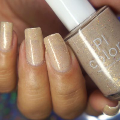 A close-up shows a hand with nails painted in a shimmery sheer beige polish, holding the bottle of Shark's Eye Moonshell .062 Nude Cream Nail Polish. The handmade bottle from PI Colors, featuring holographic glitter, is nestled between the fingers, with its distinctive brand partially visible. The background reveals a subtle, colorful bokeh effect.