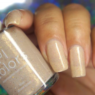 A close-up of a hand showcases neatly manicured nails painted with PI Colors' Shark's Eye Moonshell.062 Nude Cream Nail Polish, featuring a shimmery sheer beige hue. The fingers clasp a bottle of the handmade polish, which beautifully matches the nails and reveals subtle holographic glitter in the polish.