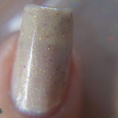 Highlighted is a fingernail adorned with PI Colors' Shark's Eye Moonshell.062 Nude Cream Nail Polish, which features a delicate shimmer in sheer beige with accents of holographic glitter. This handmade polish produces a captivating, multicolored effect as the nail is sharply focused against an elegantly blurred background.