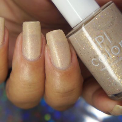 A hand with long nails painted in the shimmery sheer shade of "Shark's Eye Moonshell.062 Nude Cream Nail Polish" holds a bottle of the same artisanal polish. The bottle features a white cap, partially revealing the letters "PI Colors," adding an elegant touch to the look.