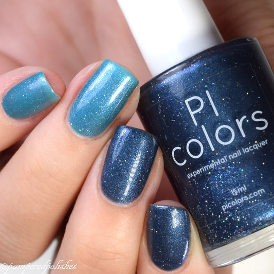 Close-up of a hand with nails painted in shimmering teal blue shades. A bottle of PI Colors' Tanager.043 Blue Nail Polish is held between the fingers, its glittery dark blue hue adding an opulent touch to your manicure.