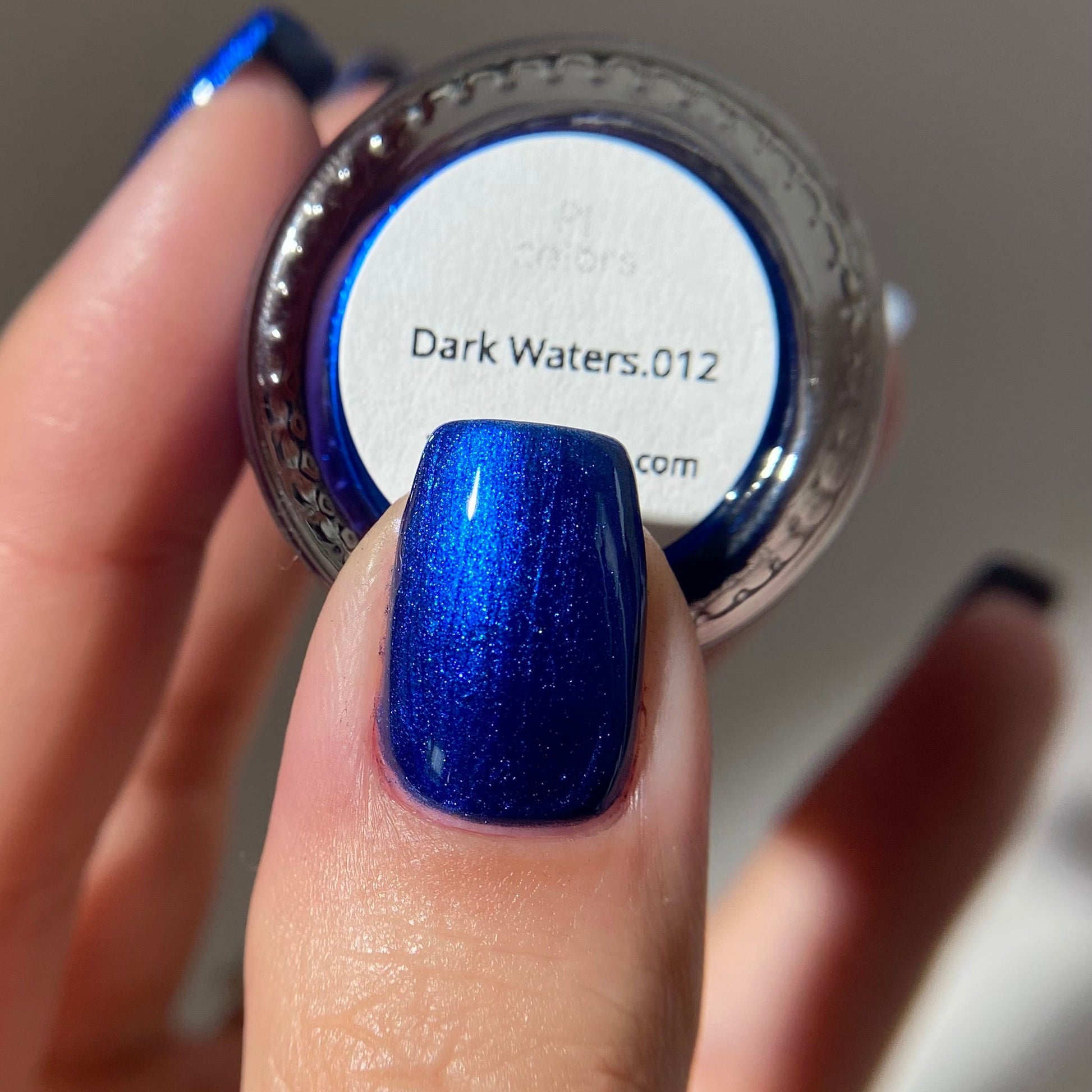 Blue glittery nail polish on a finger with a nail polish container labeled 'Dark Waters.012' in the background.