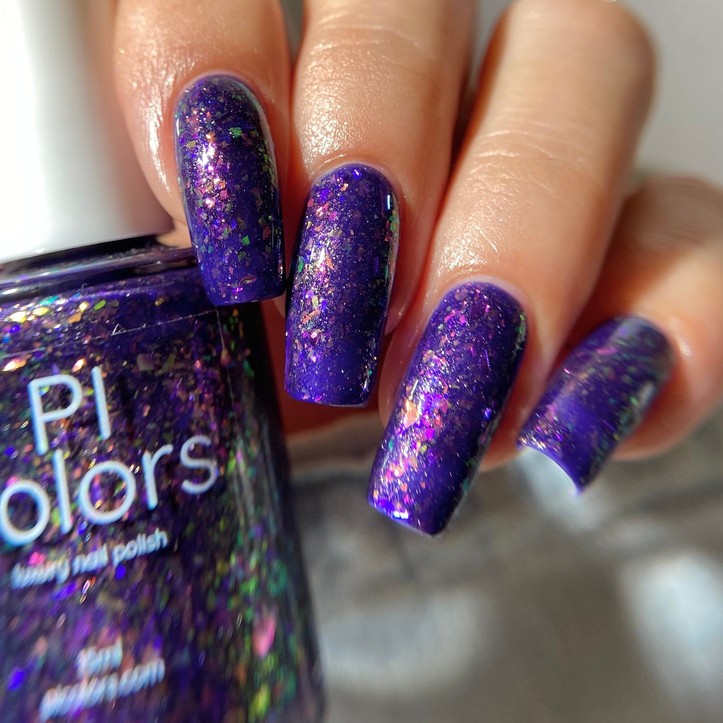 A close-up of a hand with nails painted in a deep purple holographic glitter nail polish, holding a bottle of Pi Colors Galaxy nail polish.
