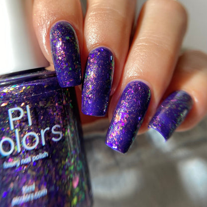 A close-up of a hand with nails painted in a deep purple holographic glitter nail polish, holding a bottle of Pi Colors Galaxy nail polish.