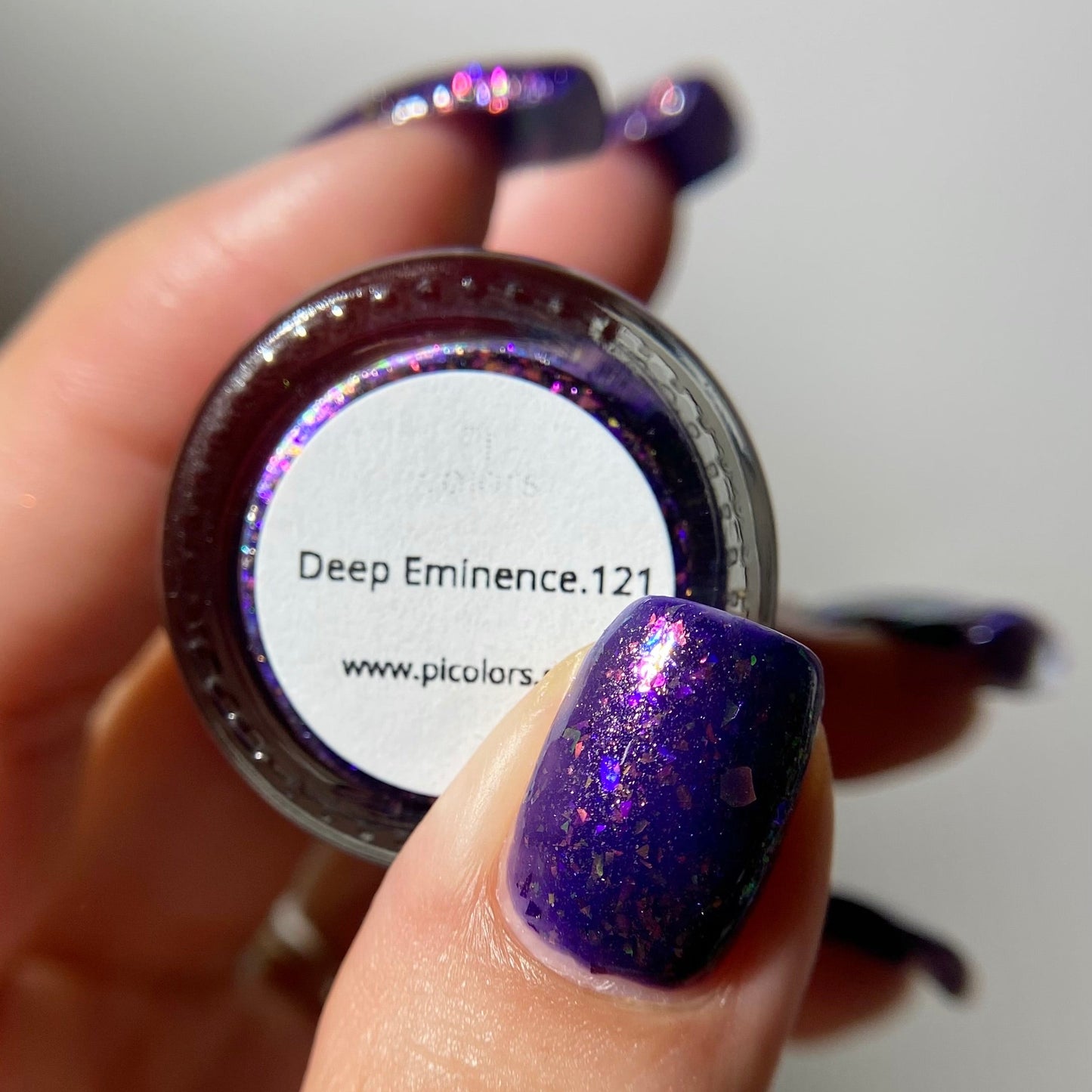 A close-up of a hand holding a small round pot of glitter nail polish. The nail polish is a deep purple with fine holographic shimmer, and the pot label reads 'Deep Eminence.