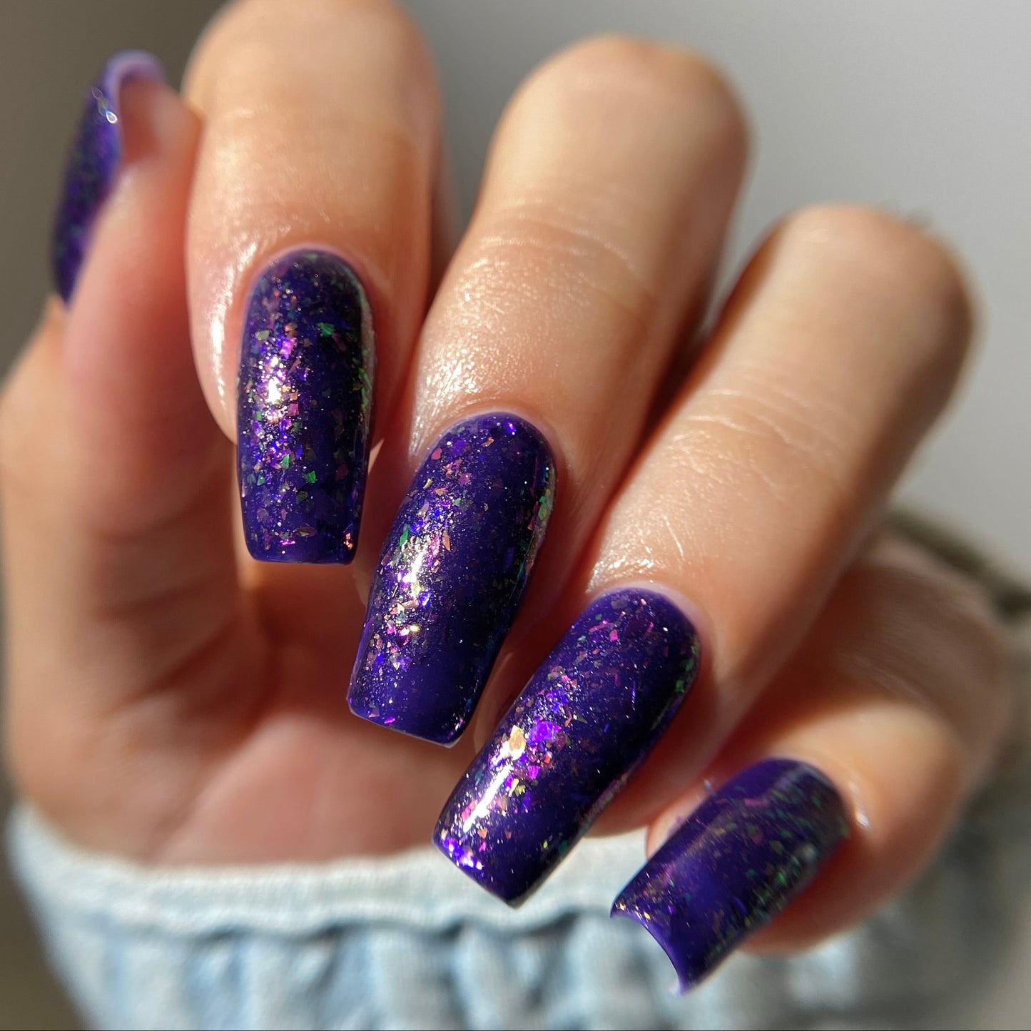 A close-up of a hand with long, almond-shaped nails painted in a deep purple glitter nail polish with fine gold shimmer, wearing a light blue knit sleeve.
