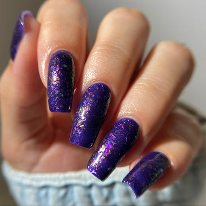 A close-up of a hand with long, almond-shaped nails painted in a deep purple glitter nail polish with fine gold shimmer, wearing a light blue knit sleeve.