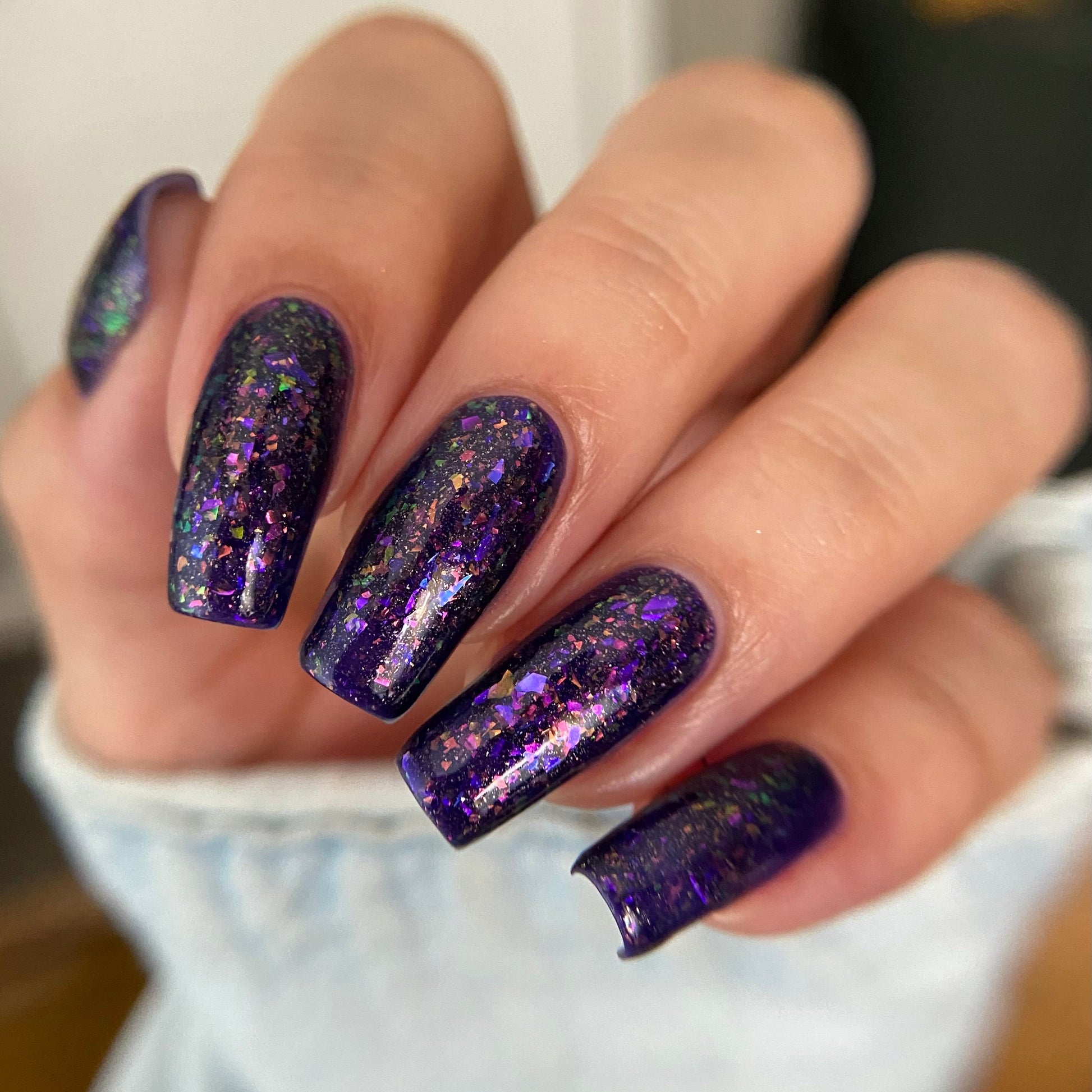 A close-up of a hand with long, almond-shaped nails painted in a deep purple glitter nail polish with iridescent flecks that shift into pink and gold, wearing a light denim jacket.
