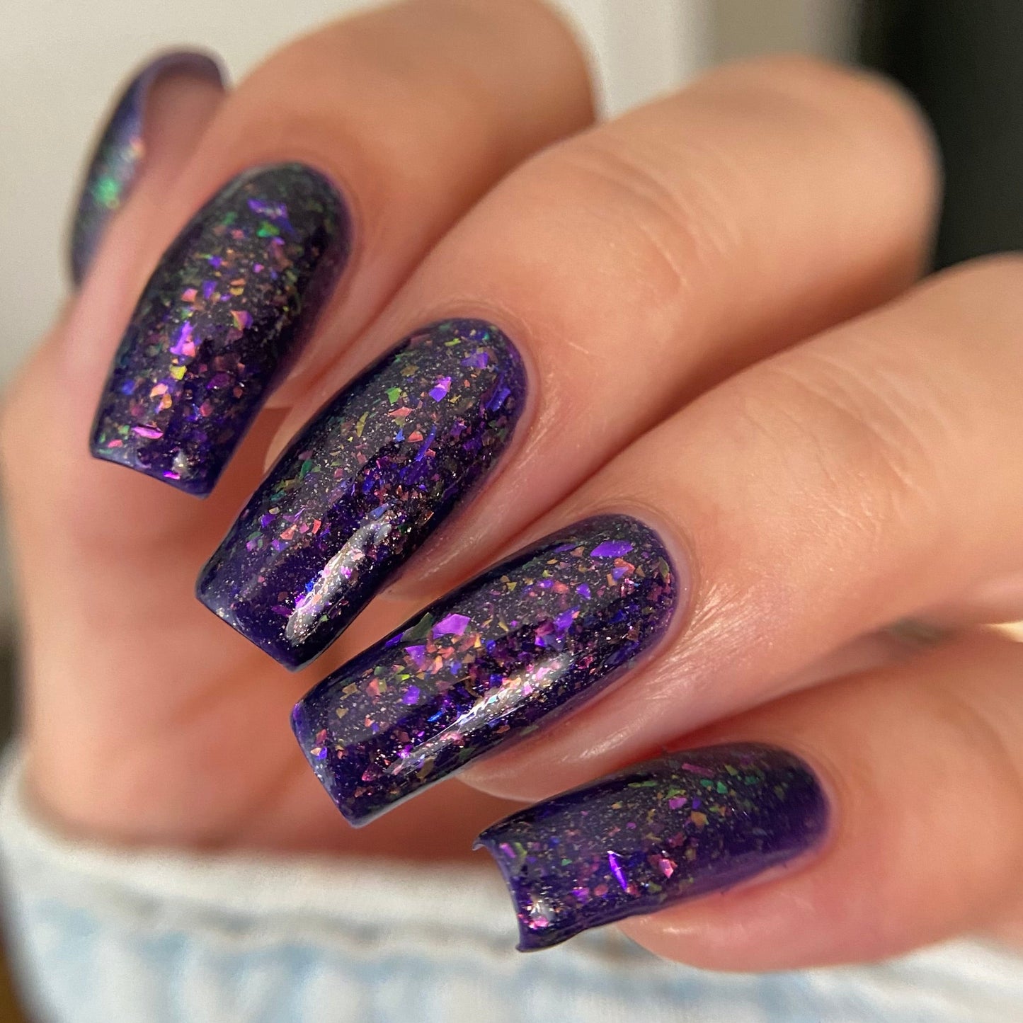 A close-up of a hand with long, almond-shaped nails painted in a deep purple glitter nail polish with fine holographic flecks.