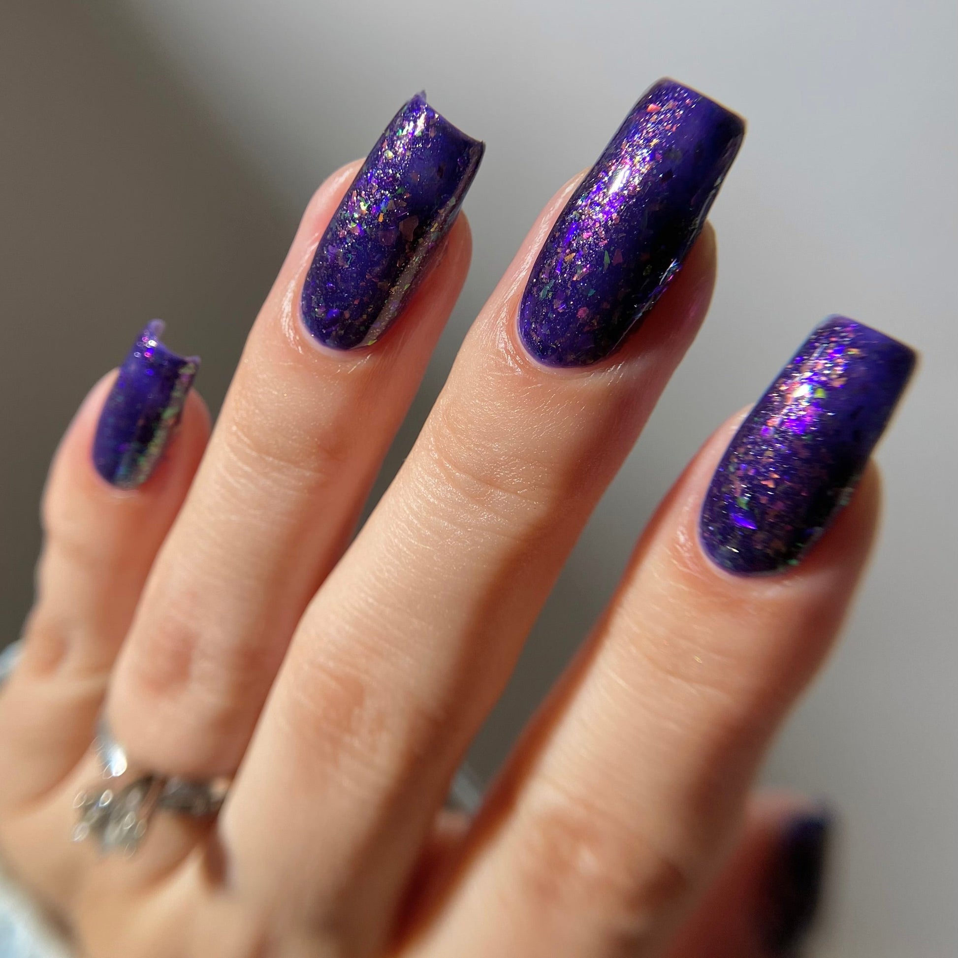 A close-up of a hand with long, almond-shaped nails painted in a deep purple glitter nail polish, wearing a silver ring on the ring finger.