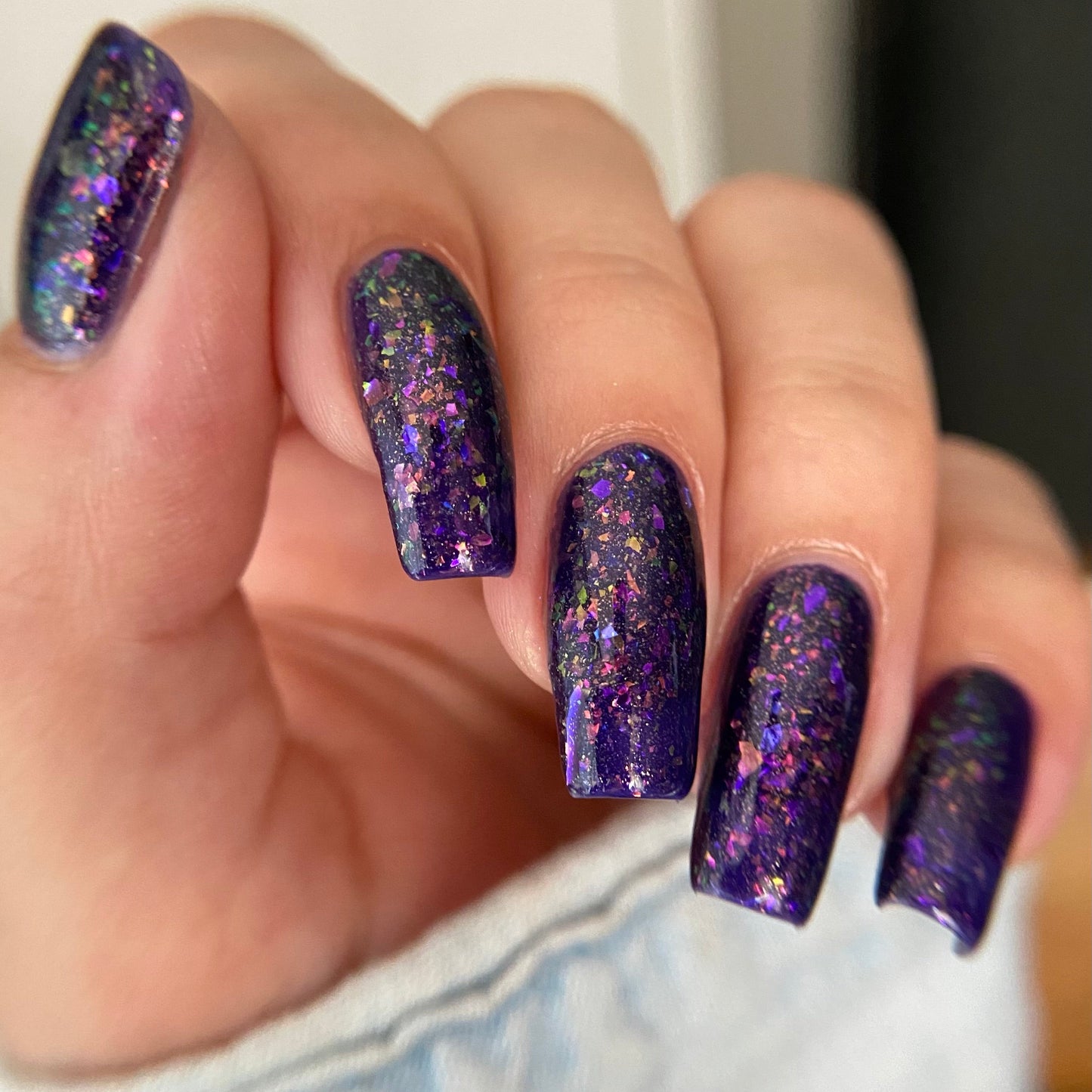 A close-up of a hand with long, almond-shaped nails painted in a deep purple glitter nail polish with iridescent flecks reflecting pink, blue, and gold.