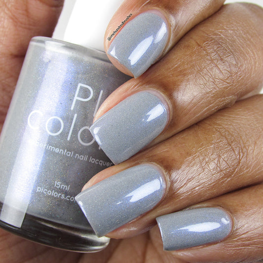 Close-up of a hand showcasing nails painted with a shimmering blue-gray nail polish accented by subtle holographic glitter. A bottle of the same handmade nail polish is held, labeled "PI Colors" and described as "Draco.055 Experimental Nail Lacquer.