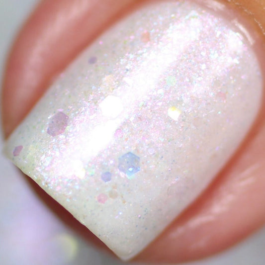 Here's a close-up of a fingernail beautifully adorned with Troth of Innocence.001 White Nail Polish from PI Colors, showcasing its handmade white crelly finish enhanced by iridescent glitter and holographic hexagonal specks. This combination results in a sparkling effect under light, offering a slightly textured and glossy look.