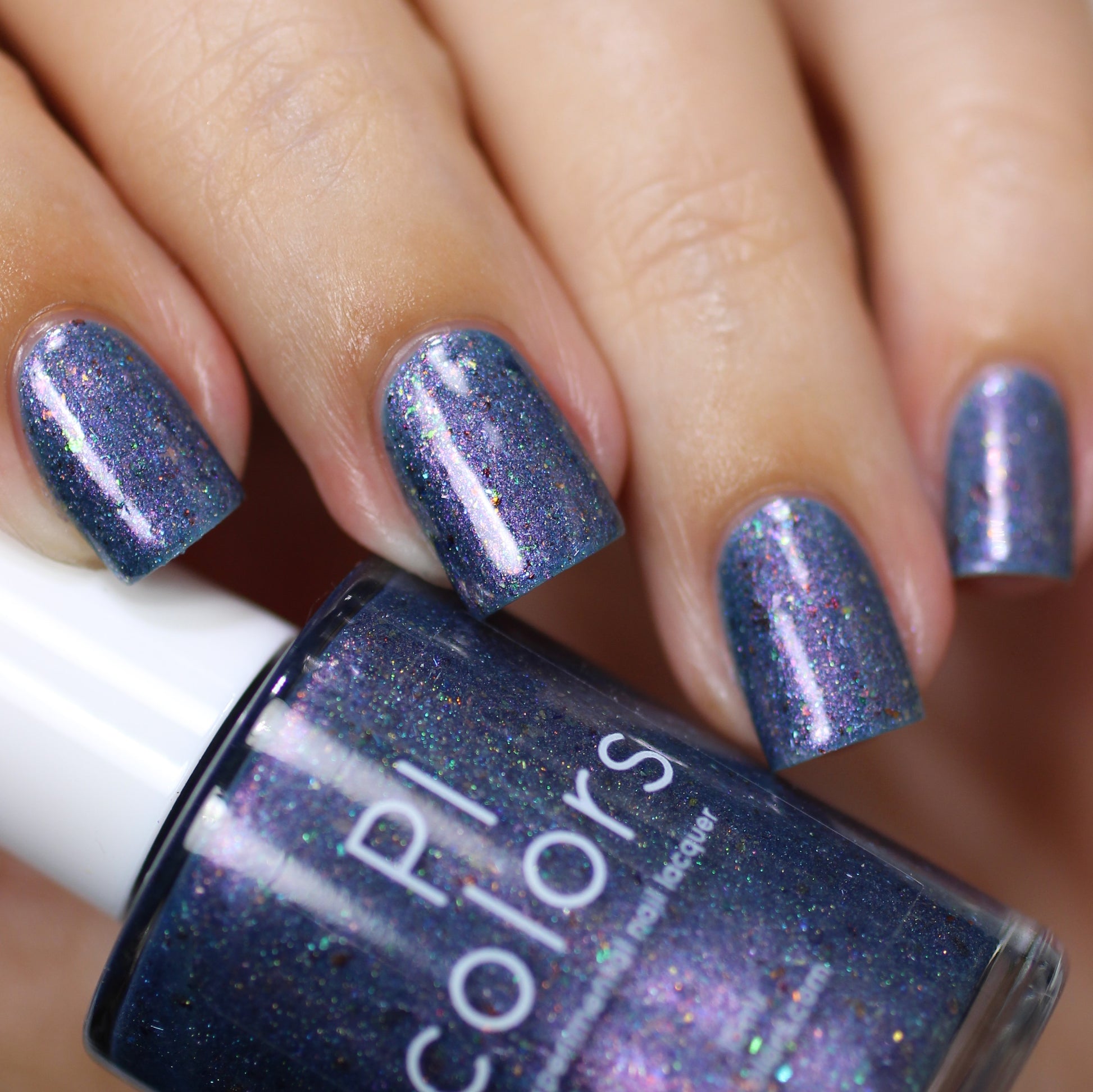 A close-up view features a hand with short nails painted in a shimmering blue-purple hue, accentuated by color-shifting iridescent flakies. In the foreground, a bottle of "East of the Storm.029 Blue Nail Polish" by PI Colors is displayed, showcasing its deep blue polish enhanced with captivating UCC flakies.