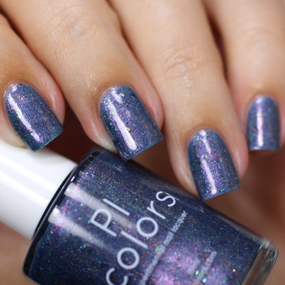 A close-up view features a hand with short nails painted in a shimmering blue-purple hue, accentuated by color-shifting iridescent flakies. In the foreground, a bottle of "East of the Storm.029 Blue Nail Polish" by PI Colors is displayed, showcasing its deep blue polish enhanced with captivating UCC flakies.