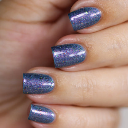 A close-up of a hand reveals nails adorned with PI Colors' East of the Storm.029 Blue Nail Polish, shimmering with flakies. The short, square-shaped nails glisten as the iridescent particles reflect a spectrum of colors, and the blurred background accentuates the vibrant allure of this stunning manicure.