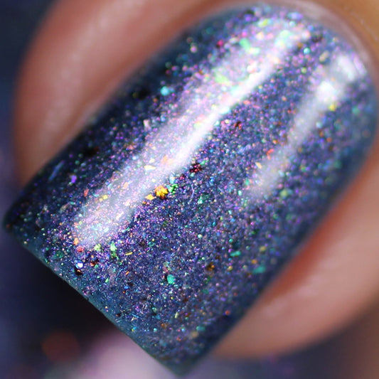 A mesmerizing close-up of a fingernail adorned with the shimmering East of the Storm.029 Blue Nail Polish by PI Colors reveals color-shifting iridescent flakes that reflect light, showcasing flecks of blue, purple, and green. The glittery surface exudes a vibrant, cosmic appearance.