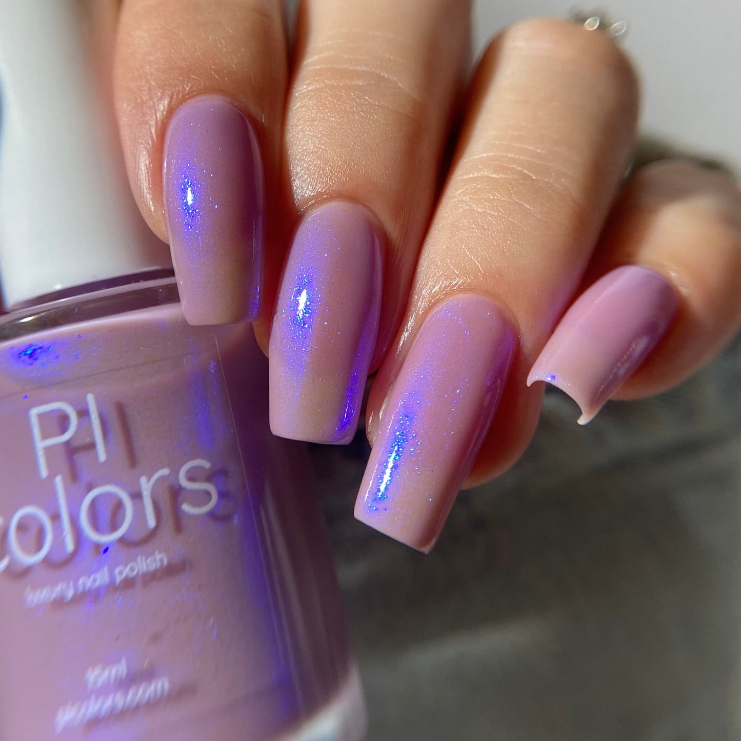 A close-up of a hand with long, square-shaped nails painted in a light lavender purple nail polish with a subtle blue shimmer, holding a bottle of Pi Colors long nail polish.