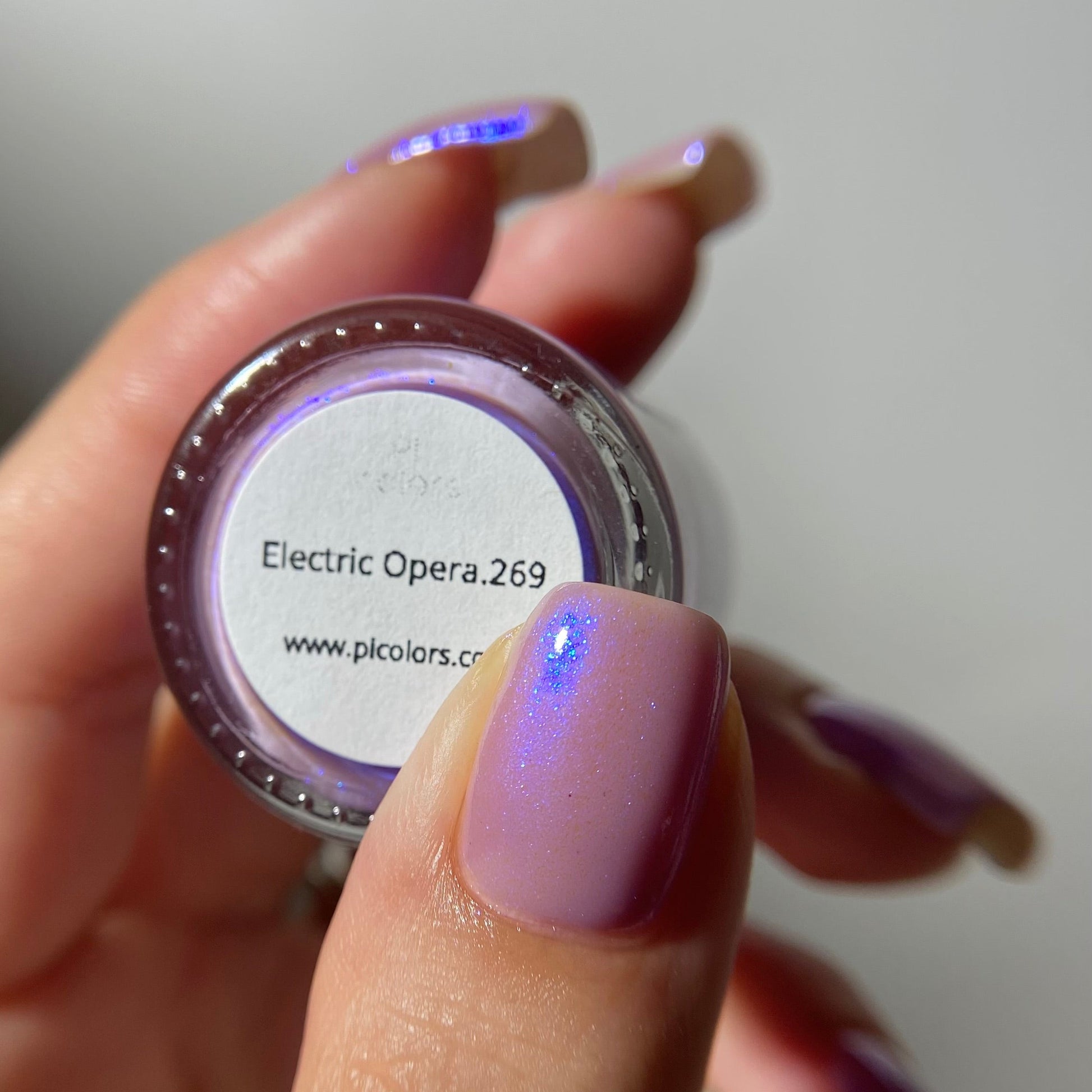 A close-up of a hand holding a small round pot of nail polish. The nail polish has a lavender-purple base with a strong electric blue iridescent shimmer.