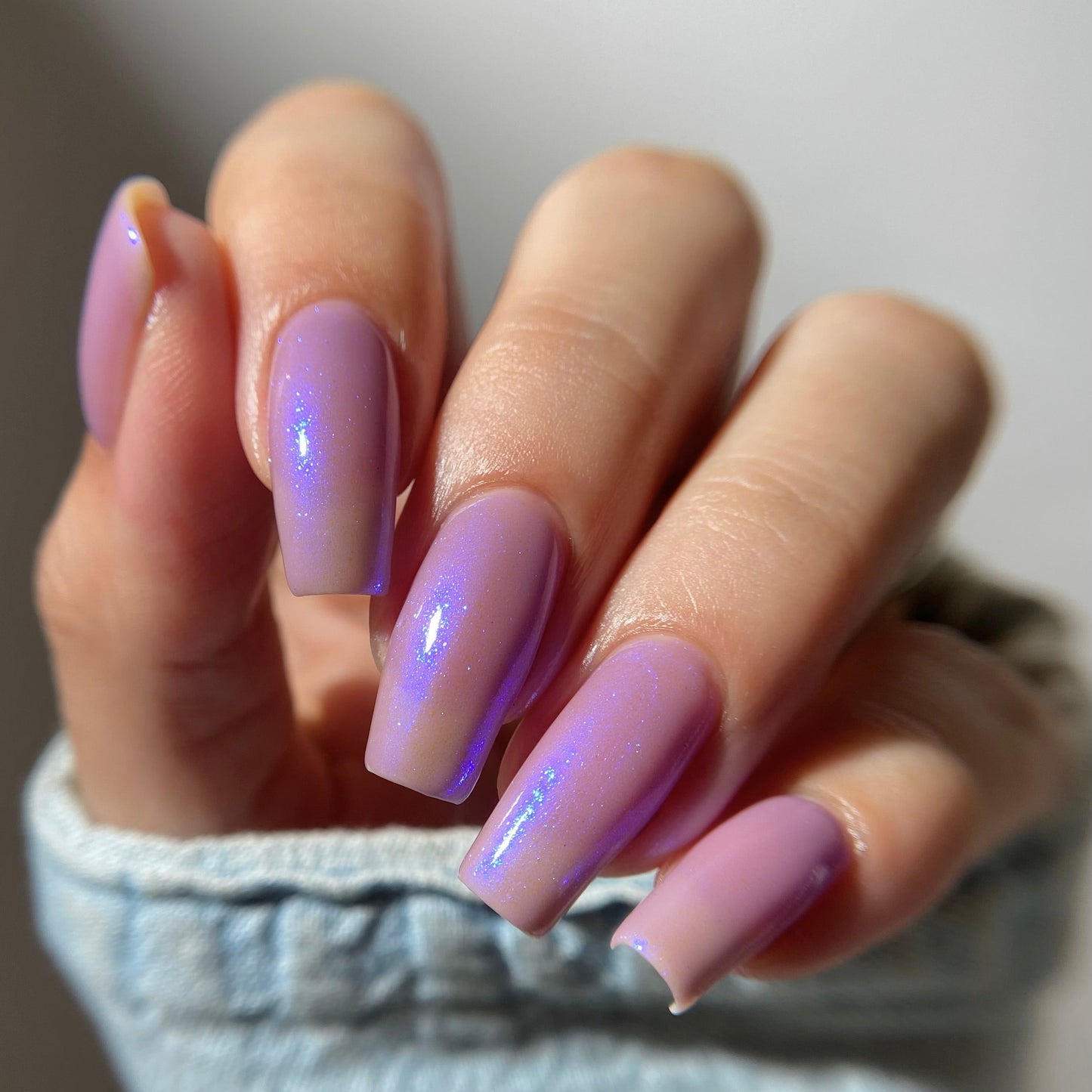 A close-up of a hand with long, almond-shaped nails painted in a glossy lavender-purple shade with a subtle iridescent shimmer, wearing a light blue denim sleeve.
