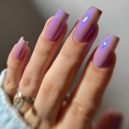 A close-up of a hand with long, almond-shaped nails painted in a glossy lavender-purple shade with fine silver glitter, wearing silver rings and a light blue knit sleeve.