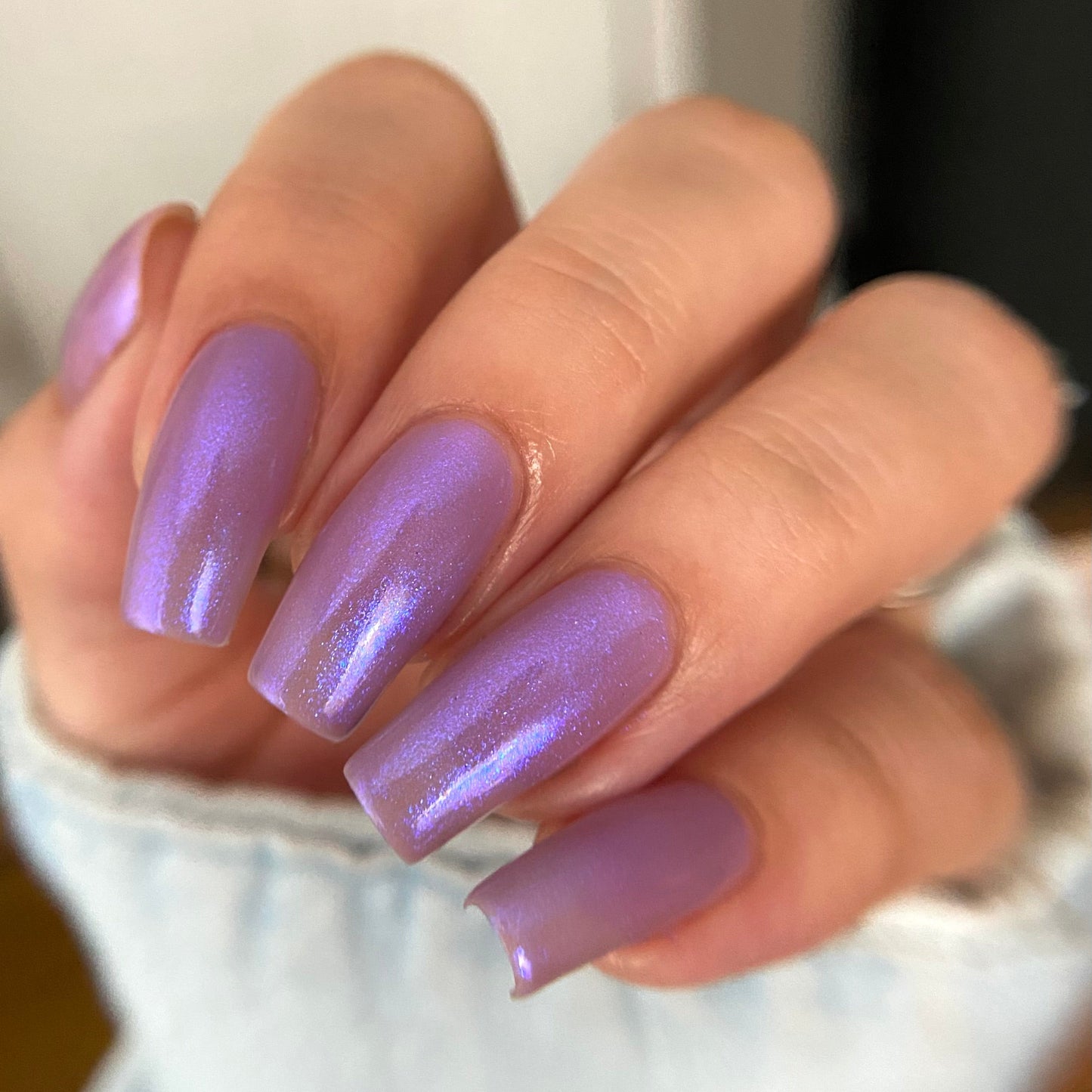 A close-up of a hand with long, almond-shaped nails painted in a glossy, shimmery lavender-purple nail polish.