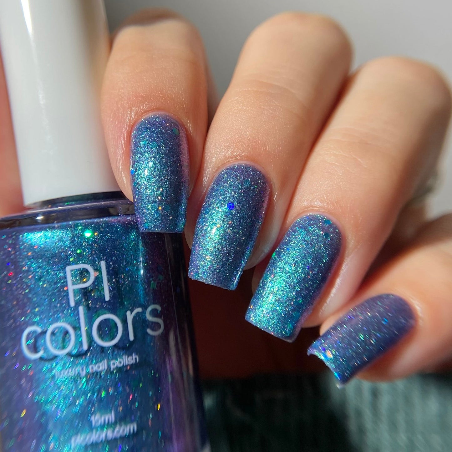 Hand with blue glittery nail polish holding a bottle of PI Colors nail polish.