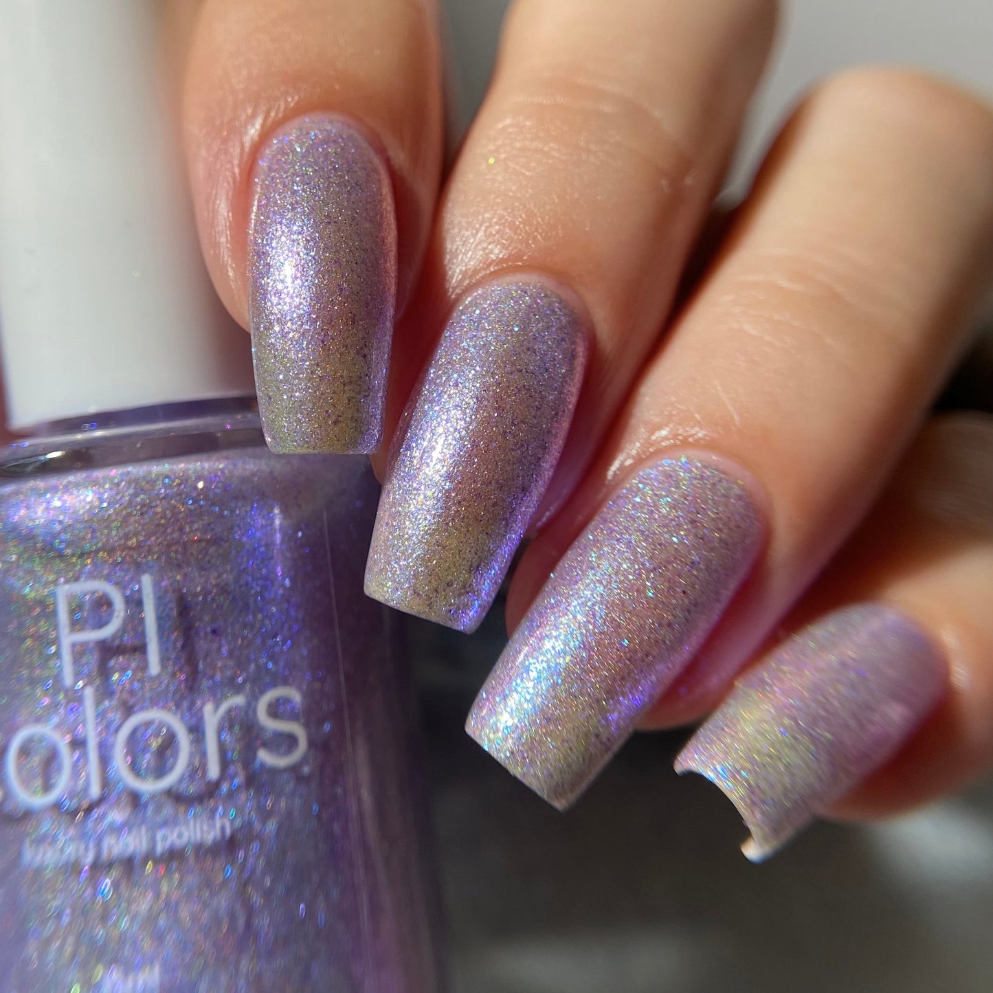 A close-up of a hand with nails painted in a shimmering lavender-purple nail polish, holding a bottle of PI Colors holographic nail polish.